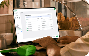 Um senhor em uma padaria mexe na Lista de Pagamento na Conta Stone pelo computador. À frente, uma maquininha da Stone e ao fundo um expositor com salgados.