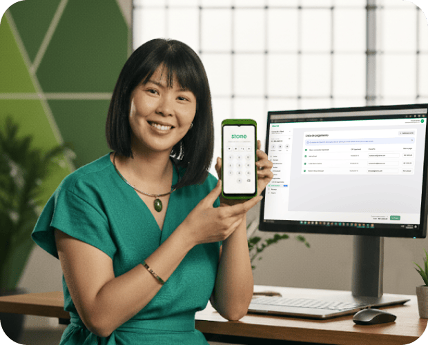 Mulher sorrindo, segurando a maquininha da Stone. Ao fundo, um computador na tela de dashboard da Conta Stone.