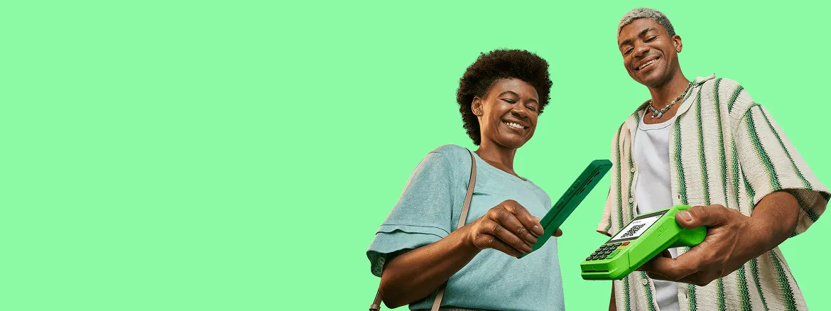 Empreendedor sorrindo, segurando a maquininha do Ton em sua mão esquerda, uma cliente está em sua frente também sorrindo e aproximando seu celular sobre a tela da maquininha para ler o QR Code que aparece na tela e realizar um pagamento via Pix.